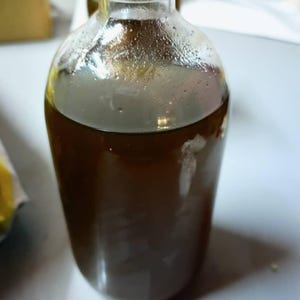 May include: A clear glass bottle filled with a dark brown, opaque liquid. The bottle has a rounded body, a narrow neck, and a white screw-top cap. The liquid inside appears viscous and fills the container nearly to the top of the neck.