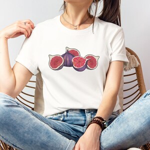 May include: White t-shirt featuring a fig illustration. The design shows whole and sliced figs in shades of purple, red, and brown. The person is wearing blue jeans and sitting in a wicker chair.