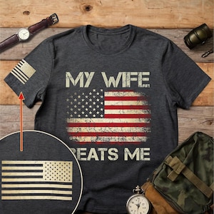 May include: Dark gray t-shirt featuring a distressed American flag design with the text "MY WIFE BEATS ME." The shirt also has a small American flag on the sleeve. A watch, a grenade, and a bag are visible.