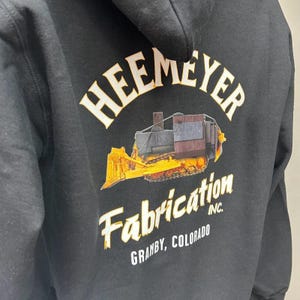 May include: Black hoodie with a white graphic of a yellow bulldozer with the text "Heemeyer Fabrication Inc. Granby, Colorado" on the back.