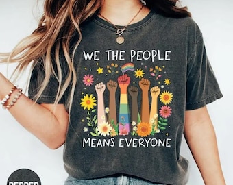 Pode incluir: T-shirt cinza escuro com a frase "WE THE PEOPLE MEANS EVERYONE". O design apresenta punhos levantados em vários tons de pele, uma bandeira arco-íris e detalhes florais. Uma mensagem de inclusão e unidade.
