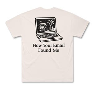 May include: Cream-colored t-shirt featuring a black graphic of a laptop displaying a beach scene with a wave, palm tree, and surfboard. The text "How Your Email Found Me" is printed below the laptop image. The shirt has short sleeves and a crew neck.