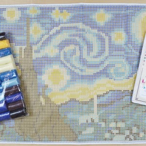 May include: A latch hook kit featuring a blue and yellow starry night scene. The kit includes a canvas, yarn, and a hook. The instructions are printed on a separate sheet of paper.