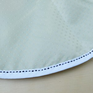 May include: Close-up of a light green fabric with a white trim and a black stitching detail. The fabric has a subtle dotted pattern.