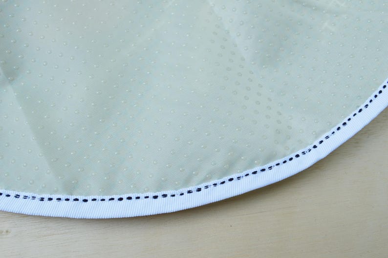 May include: Close-up of a light green fabric with a white trim and a black stitching detail. The fabric has a subtle dotted pattern.