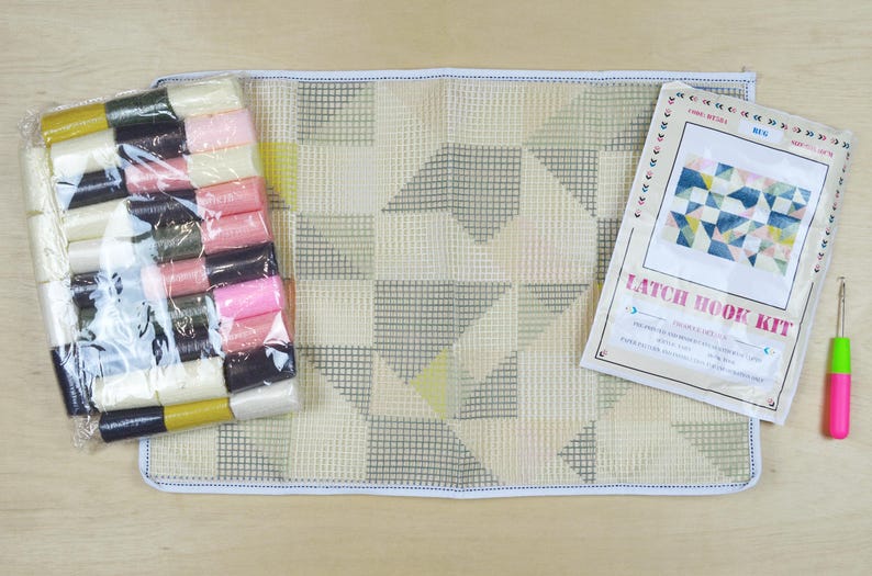 May include: A latch hook kit with a white canvas, colourful yarn, a hook, and instructions. The design features a geometric pattern of triangles and squares in shades of blue, pink, and yellow. The instructions include the text "Latch Hook Kit".