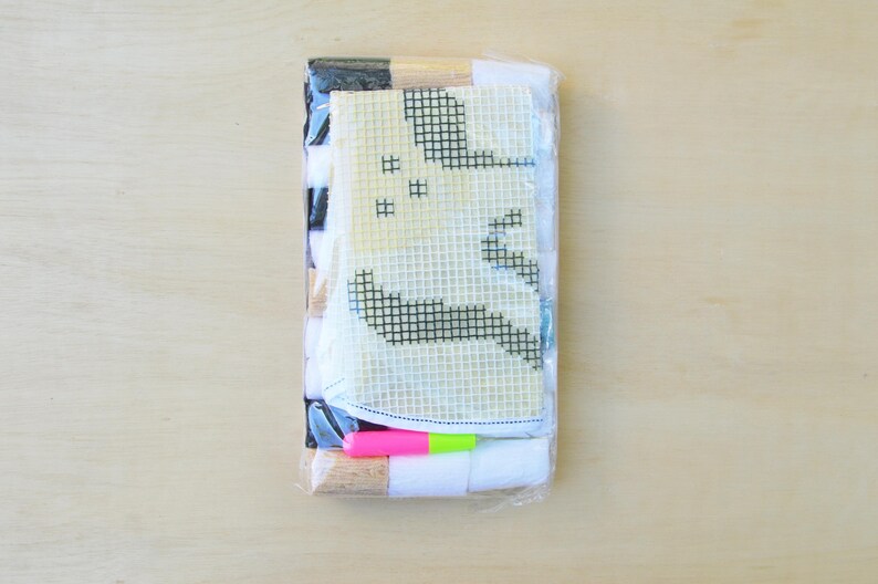 May include: A plastic-wrapped kit for creating a cross-stitch project. The kit includes a white canvas with a black and white design of a ghost, colourful threads, and a wooden needle.