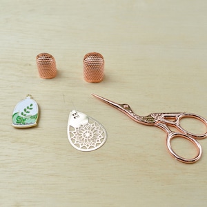 May include: A set of sewing tools including two rose gold thimbles, a rose gold pair of scissors with a bird design, a rose gold needle threader, and a small enamel charm with a green plant inside a glass dome.