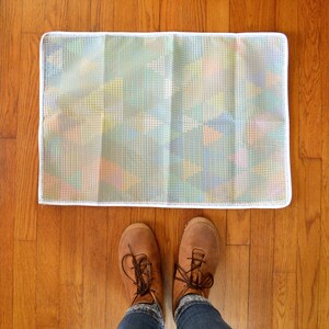 May include: A white doormat with an iridescent, grid-like pattern. The doormat is on a wooden floor.