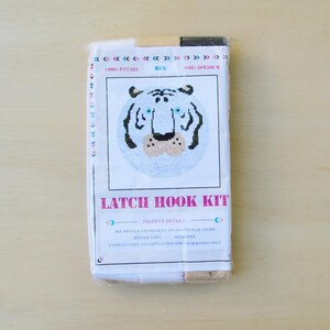May include: A latch hook kit featuring a white tiger face with black stripes and blue eyes. The kit includes yarn, canvas, and instructions. The packaging is white with pink text that reads "Latch Hook Kit".