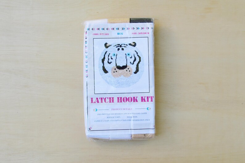 May include: A latch hook kit featuring a white tiger face with black stripes and blue eyes. The kit includes yarn, canvas, and instructions. The packaging is white with pink text that reads "Latch Hook Kit".
