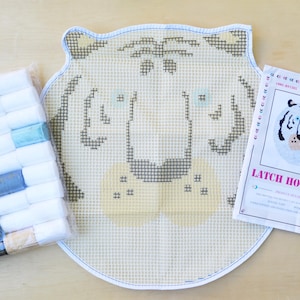 May include: A latch hook kit featuring a white tiger face design. The kit includes a canvas, yarn, and a hook. The canvas is white with a grid pattern and the tiger face is outlined in black. The yarn is in a variety of colours, including white, black, and orange. The hook is pink and has a metal hook.