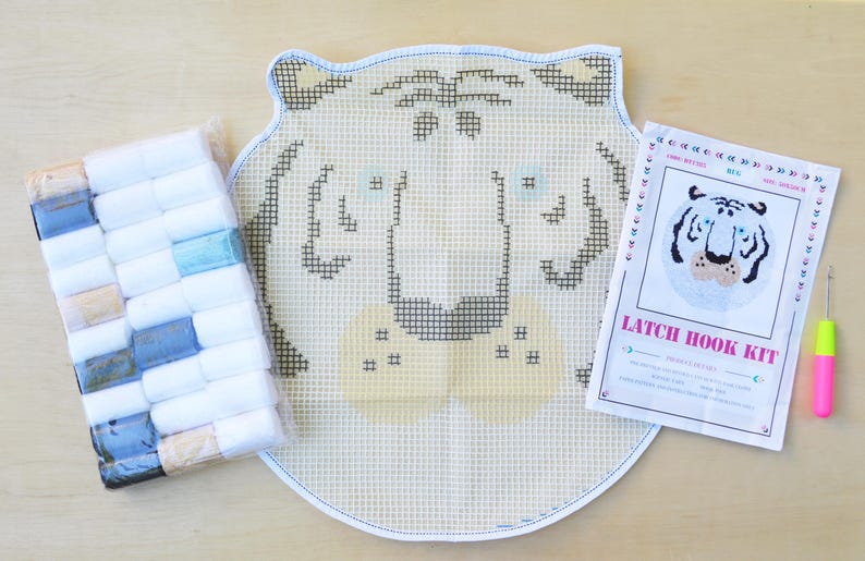 May include: A latch hook kit featuring a white tiger face design. The kit includes a canvas, yarn, and a hook. The canvas is white with a grid pattern and the tiger face is outlined in black. The yarn is in a variety of colours, including white, black, and orange. The hook is pink and has a metal hook.