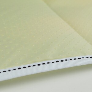 May include: Close-up of a light yellow fabric with a white trim and black stitching. The fabric has a subtle dotted pattern.