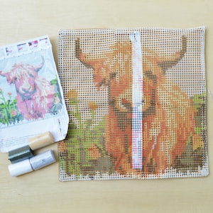 May include: A close-up of a needlepoint canvas with a design of a brown and white Highland cow. The canvas is white with a grid pattern. There are also some yarn and a needlepoint needle in the image.