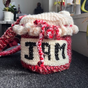 Op de afbeelding: Een gehaakte rode en witte tas in de vorm van een pot met een wit gehaakt deksel en een zwart gehaakt trekkoord. De tas heeft het woord "JAM" in zwart garen geborduurd op de voorkant.