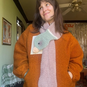 May include: A handmade duck scarf with a light green head and a pink body. The scarf has crocheted feet and a beak. The person in the image is wearing an orange fleece jacket.