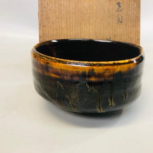 May include: A ceramic tea bowl with a dark interior and a textured exterior. The rim is a warm, golden-brown color, transitioning to a deep, earthy brown. A wooden box is in the background.