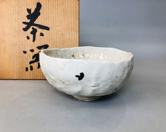 May include: A white ceramic tea bowl with a rough, textured surface. The bowl has a slightly irregular shape and is decorated with small black and green spots. The bowl is sitting on a wooden surface.