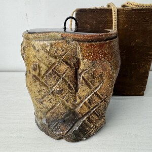 Può includere: Un vaso in ceramica cilindrico e testurizzato con una smaltatura maculata marrone e marrone chiaro, caratterizzato da un profondo motivo a tratteggio incrociato sull'esterno. Il vaso ha un coperchio scuro, liscio e piatto con una piccola maniglia in metallo nero.