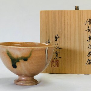 May include: A peach-coloured ceramic tea bowl with a footed base, decorated with dark green and black accents. A wooden box with Japanese calligraphy is behind the bowl.