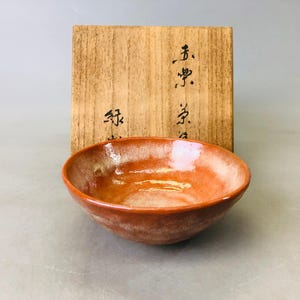 Könnte beinhalten: Handgefertigte, orange-braune Keramikschale mit glänzendem Finish. Die Schale steht vor einer Holzkiste mit japanischer Kalligraphie. Das Innere der Schale hat ein helleres, gesprenkeltes Aussehen.