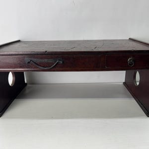May include: A dark brown wooden desk organizer featuring a rectangular top surface, one wide drawer with a curved metal handle, and one smaller drawer with a circular metal pull. The side supports have circular cutouts on each end.