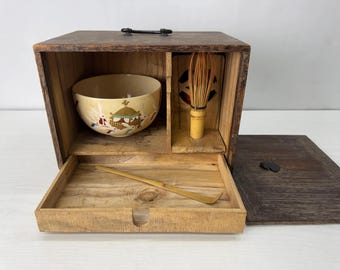 Op de afbeelding: Een houten opbergdoos voor de theeceremonie met een donkerbruine afwerking en handvat. Binnenin staat een crèmekleurige keramische kom met konijnenillustraties naast een bamboe garde. Een bamboe theeschepje ligt in de open onderste lade.