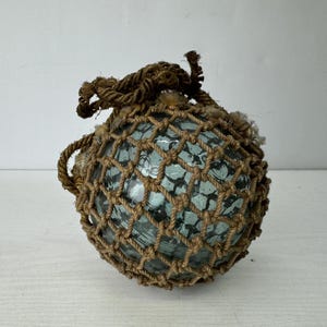 May include: A spherical, translucent light blue glass fishing float encased in a decorative, hand-knotted brown jute rope netting. The glass surface features a textured, bubbly appearance, and the rope forms a loop at the top for hanging.