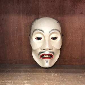 May include: A white, carved wooden mask with a detailed face. The mask features black eyebrows, mustache, and goatee, with red lips and dark eyes. The mask is set against a wooden background.