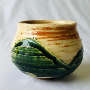 Può includere: Una piccola ciotola rotonda in ceramica con una base color crema e una glassa verde e marrone. La glassa ha un design striato e astratto.