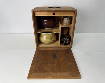 Op de afbeelding: Een houten theeceremoniedoos met een neerklapbaar voorpaneel, met daarin een donkerrode kom, een lichtgele kom, een kleine cilindrische houder, een bamboe garde en een zwarte kom met bamboepatronen, gerangschikt op interne planken.