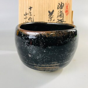 May include: A black ceramic tea bowl with a speckled glaze and a brown rim. The bowl is sitting on a wooden surface. The text "油滴" is written on the box.
