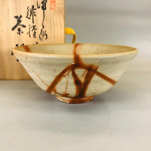 以下が含まれることがあります： 茶碗は白磁でできており、茶色の釉薬が施されています。茶碗は丸みを帯びた形をしていて、縁が少し広がっています。茶碗は木製の台の上に置かれています。