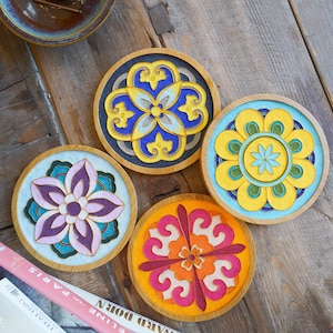 May include: Four decorative coasters with floral designs. Each coaster features a wooden frame, a colorful floral pattern, and a light blue or black background. The coasters are arranged on a wooden surface.