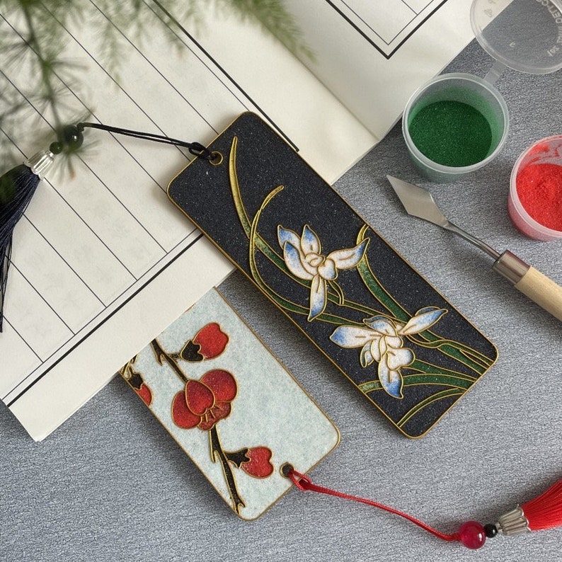 May include: Two decorative bookmarks with floral designs. One bookmark features a black background with white and blue flowers and green leaves. The other bookmark has a white background with red flowers and green leaves.