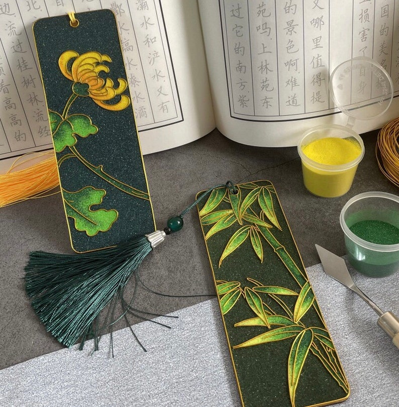 May include: Two bookmarks with green and gold designs. One bookmark features a chrysanthemum flower and the other features bamboo. Both bookmarks have a green tassel.