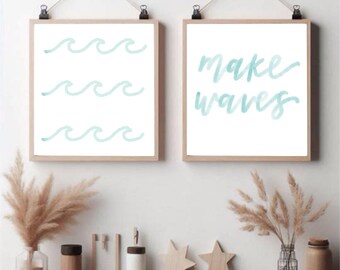 May include: Two framed prints with a beach theme. One print features three rows of watercolor waves in light blue. The other print says "make waves" in light blue script. The frames are light wood, and the prints are hung with clips.