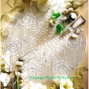 Könnte beinhalten: Eine weiße Häkeldeckchen mit einem zarten Blumenmuster. Die Deckchen liegt auf einer Holzoberfläche mit weißen Blumen, grünen Stielen und kleinen weißen Steinen.