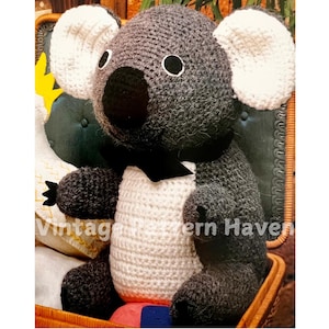 May include: A crocheted koala bear toy with gray fur, white ears, and a black bow tie. The toy is sitting in a wicker basket.
