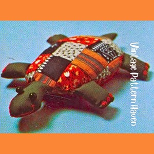 May include: A handmade turtle plushie with a patchwork shell in red, black, orange, and white patterns. The turtle has green flippers, a head, and small black eyes. The text "Vintage Pattern Haven" is visible on the right side.