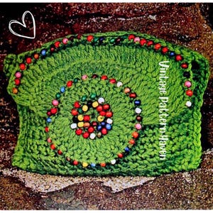 May include: A green crocheted pillow with a spiral design of colorful beads. The pillow has a square shape and is decorated with a border of beads. The text "Vintage Pattern Haven" is visible on the side.