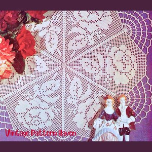 May include: A white, crocheted doily with a detailed floral pattern, displayed on a purple surface. Red roses are visible in the upper left corner. Two porcelain figurines are in the lower right corner. The text "Vintage Pattern Haven" is in the lower left corner.