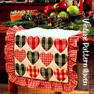 May include: A white table runner featuring a pattern of red and green plaid hearts. The runner has a ruffled edge and is decorated with fruit and greenery. The text "Vintage Pattern Haven" is visible on the side.