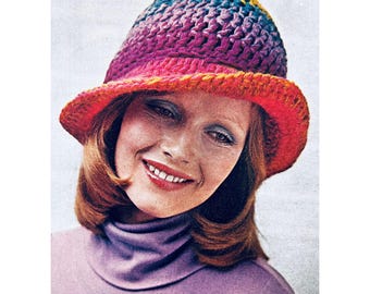 May include: A woman wearing a crocheted hat with a rainbow ombre design. The hat has a wide brim and is made with a variety of colors, including yellow, orange, red, pink, purple, and blue.