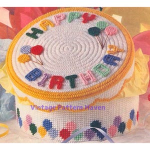 May include: A white crocheted birthday cake box with a yellow border. The top of the box is decorated with colorful letters spelling "Happy Birthday" and balloons. The bottom of the box is decorated with colorful balloons.