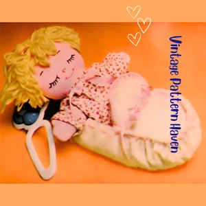 May include: A vintage doll with blonde yarn hair, closed eyes, and a smiling face, dressed in a floral print outfit, resting in a cream-colored cradle. The image includes the text "Vintage Pattern Haven" and three heart illustrations.