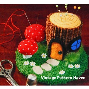 May include: A felt pincushion shaped like a tree stump with a small door and window. The stump is surrounded by green felt grass, two red mushrooms with white polka dots, and white felt flowers. The pincushion has several pins stuck in it.
