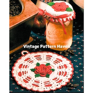 Può includere: Un sottopentola all'uncinetto rosso e bianco con una rosa rossa al centro. Il sottopentola ha un anello per appenderlo. Il sottopentola è su una superficie con motivo blu e bianco.