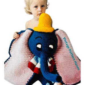 Może przedstawiać: Kocyk w kształcie słonia wykonany na szydełku z żółtym kapeluszem, niebieskim ciałem i różowymi uszami. Kocyk trzyma małe dziecko.